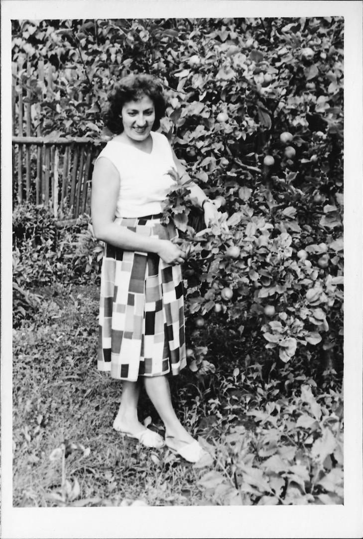 ena s po ramena dlouhmi vlnitmi vlasy v rozepnutm svetku a sukni s nepravidelnm obdlnkovm barevnm
  potiskem stoj na zahrad vedle jablon. Za n je plakov plot.