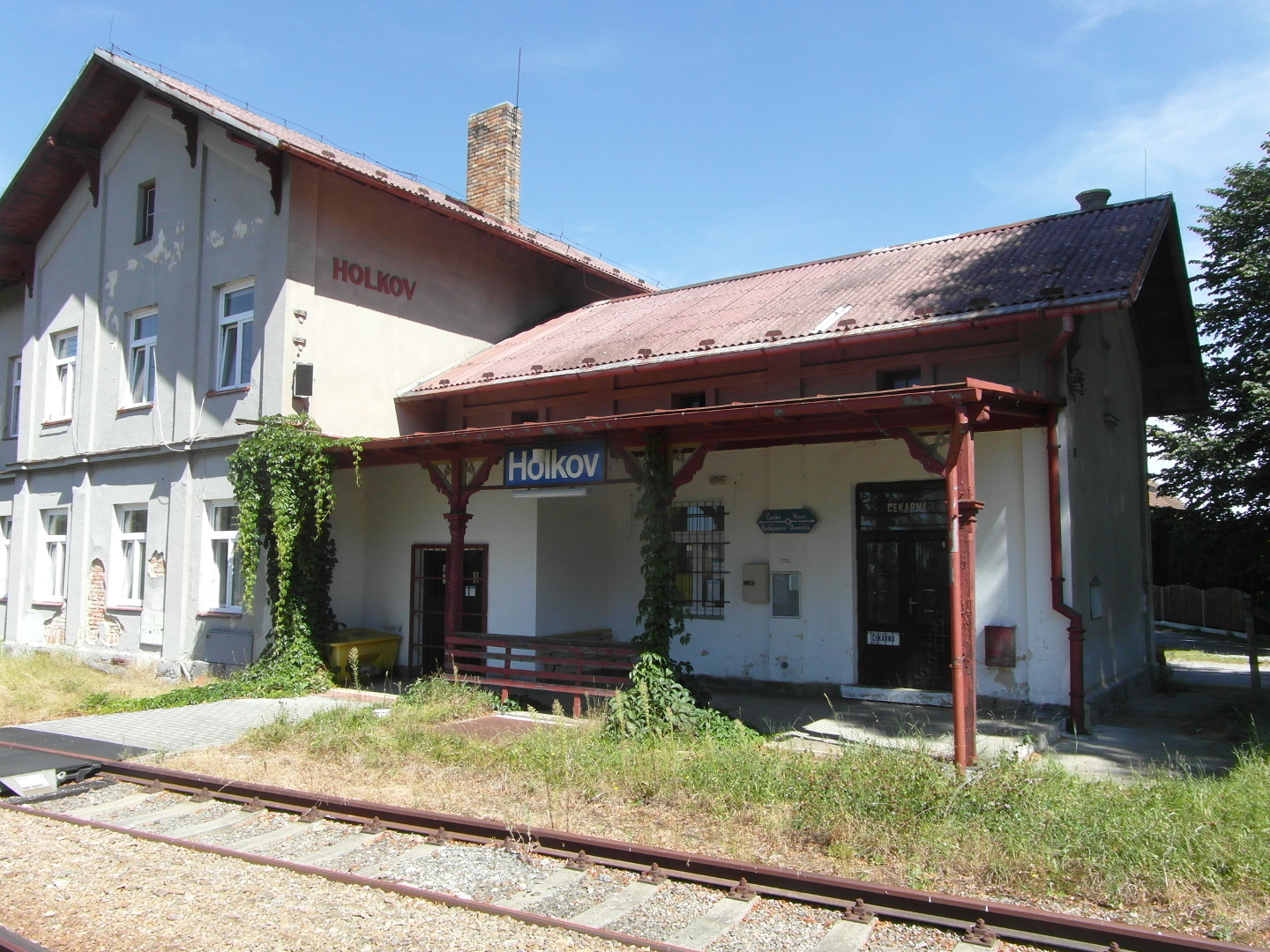 Na fotografii eleznin stanice Holkov je vidt hlavn prel, budovu s pstekem i jedny koleje. Budova je omtnuta na edo. Historick
  nzev stanice na zdi je erven.