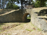 Na fotografii je kamenn most pes potok. Na most je devn zbradl a za mostem stromy.