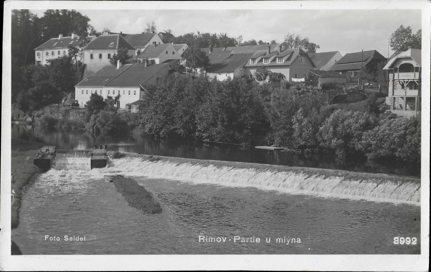 Na pohlednici je jez na ece Mali. V pozad jsou stromy na Felixov nbe. Nad nimi jsou na svahu budovy.