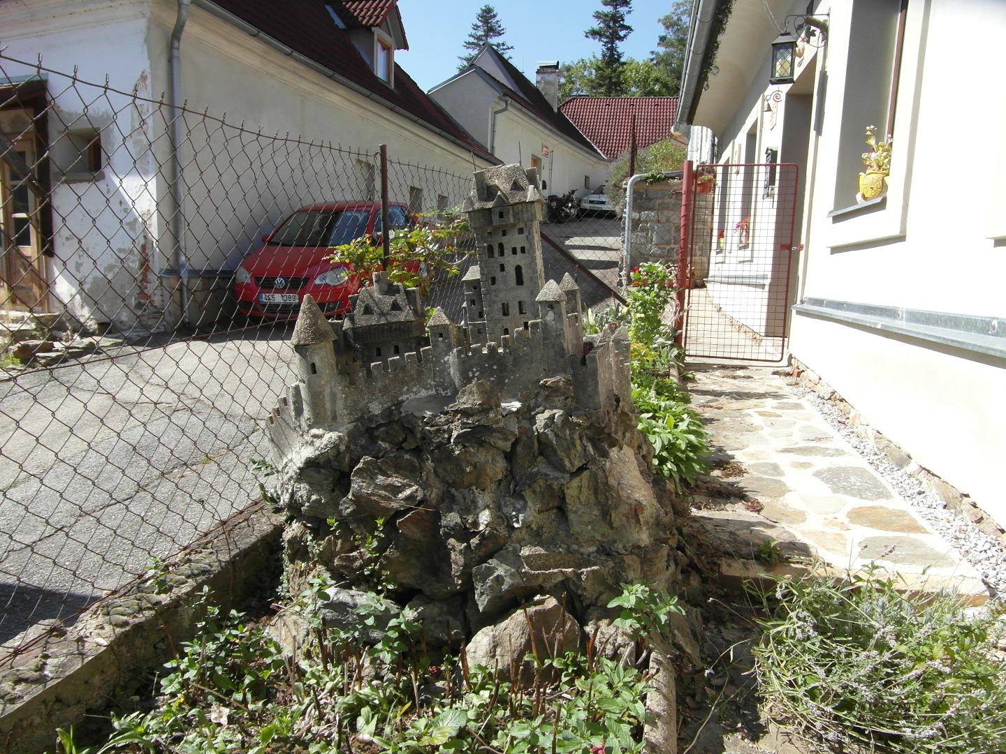 Pohled na miniaturu hradu proti svahu ulice. Dle je vidt asfaltov silnice, kameny vydldn plocha mezi zahrdkou a domem, k nmu miniatura pat a erven branka. Domy,
  kter jsou v zbru na tuto st ulice, jsou bl.     
  Miniatura hradu m vysokou hlavn v se sedlovou stechou. Tm pipomn Karltejn. Na stavbice se nachz jet palce, mnoho mench v pospojovanch hradbami s cimbum a velk
  mnostv oken nejen na palcch ale i na hlavn vi jsou otvory do interir. Pes interiry je pod uritmi hly vidt na druhou stranu. Stechy jsou opateny viki. Miniatura m
  barvu betonu. Z tohoto smru je vidt i bortc se skalka a jak moc je miniatura naklonna smrem do ulice. Na hlavn vi je sten ve stee arkek. Na stechch jsou komny
  a rezav ocelov drtky, na kterch byly praporky.