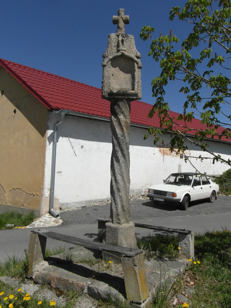 Na fotografii z vletu jsou kamenn sloupov Bo muka s ohrdkou,
            silnice, zaparkovan bl automobil, silnice a budova. Hlavice
            Boch muk je umstna na korintskm sloupu.